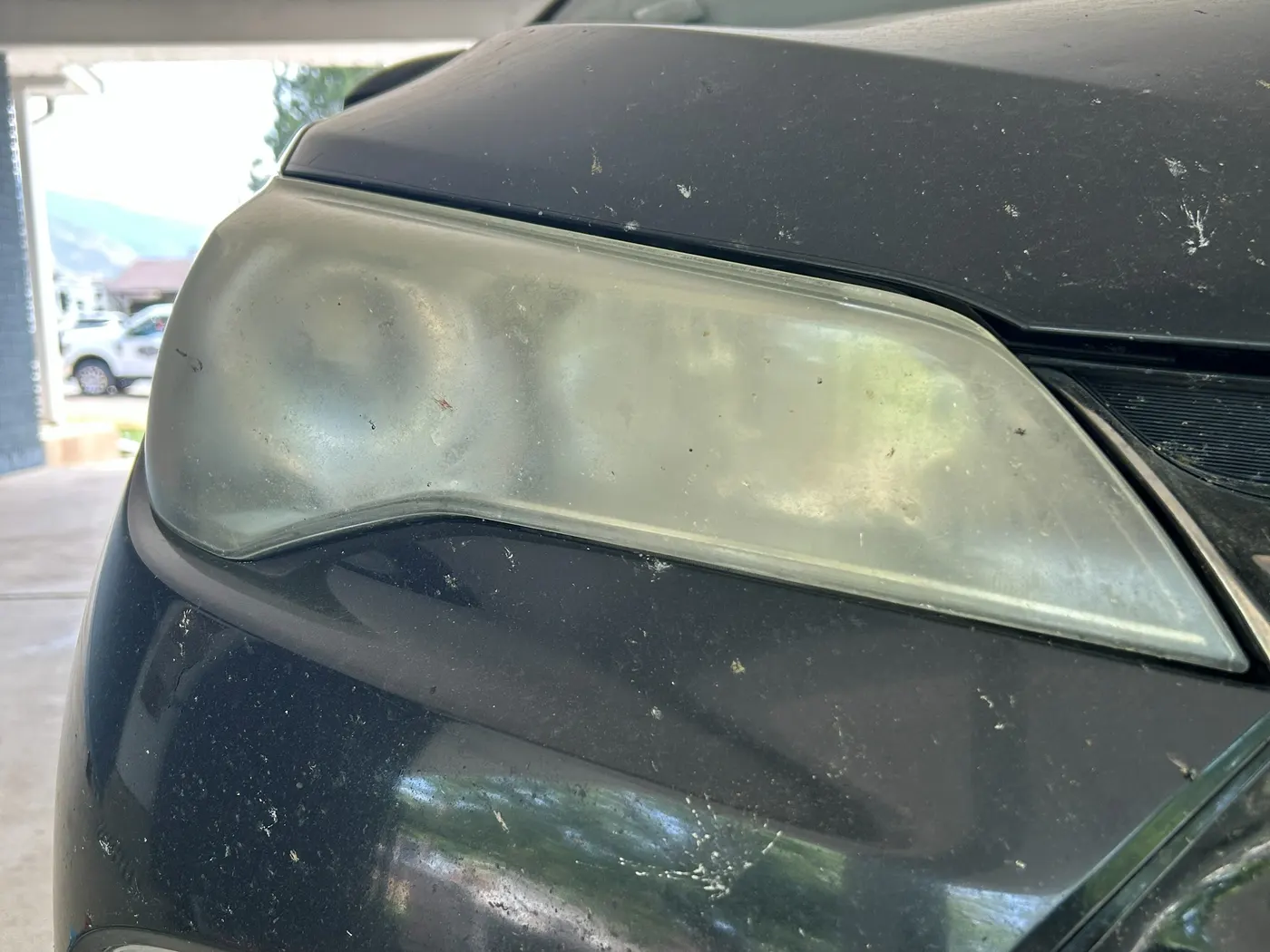 Passenger-side headlight on a black Toyota Camry before restoration, the lens clouded milky-yellow so the bulb inside is barely visible