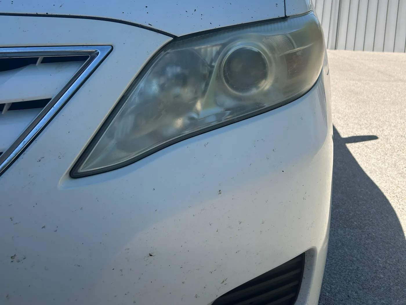 Driver-side headlight on a white Toyota Camry before restoration, shot at an angle, the lens fogged yellow with the reflector barely visible