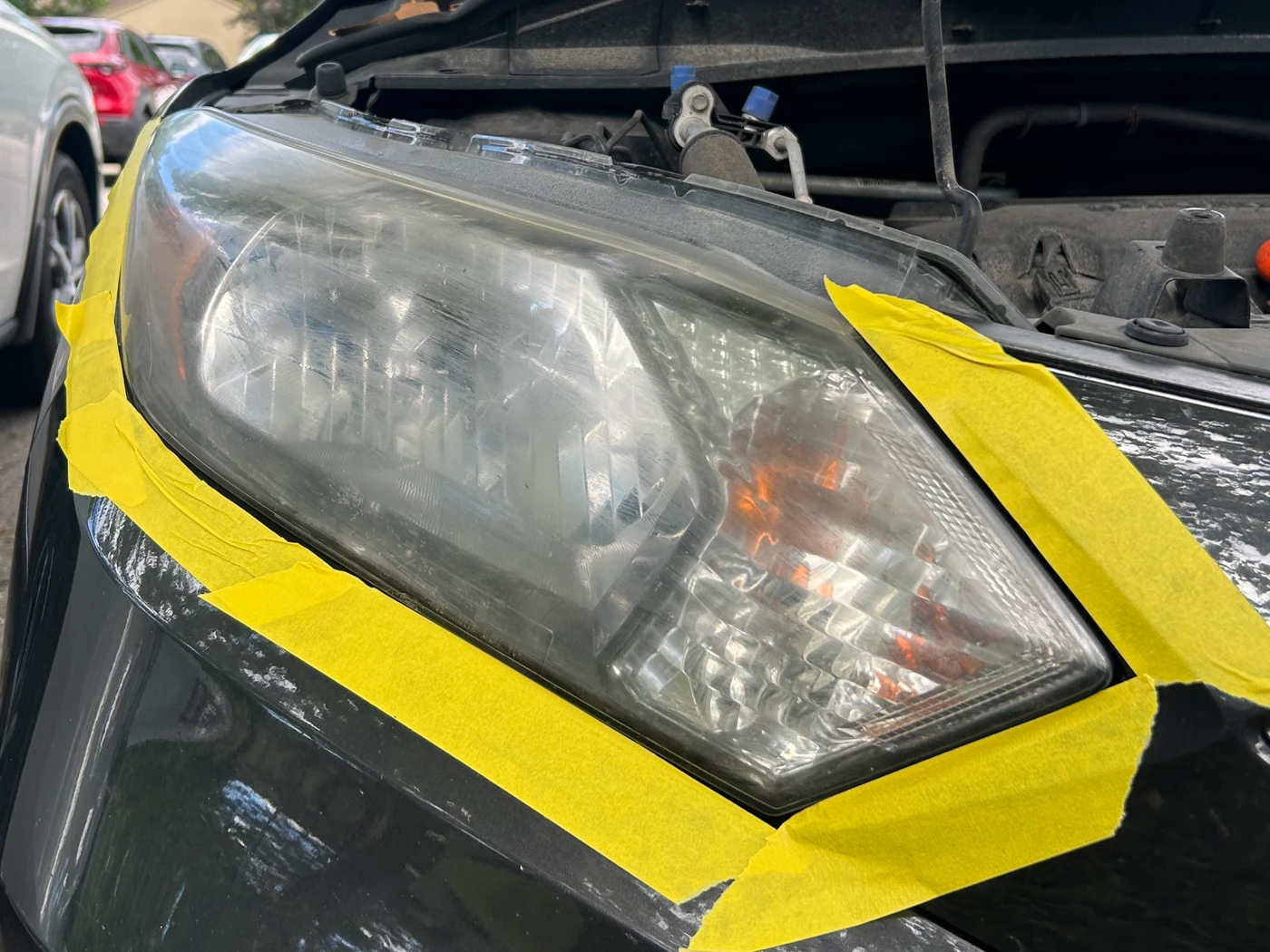 Headlight on a dark Hyundai masked off with tape before restoration, the lens clouded and hazy