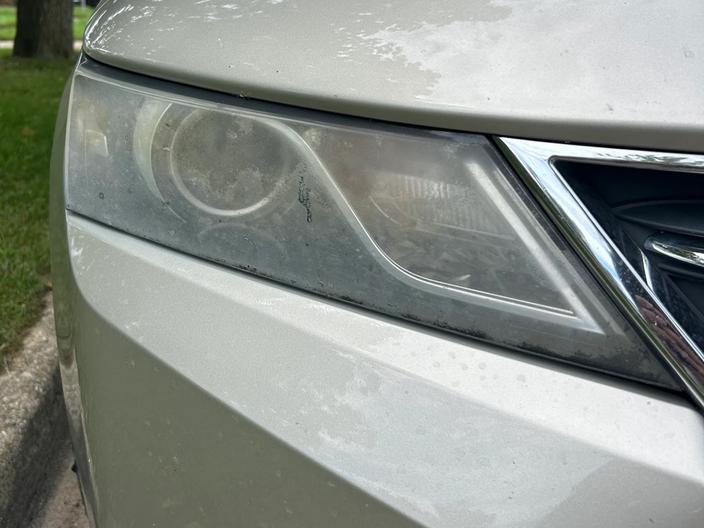 Headlight on a champagne Chevrolet Impala before restoration, the lens crusted grey-brown with oxidation so the projector inside is barely visible
