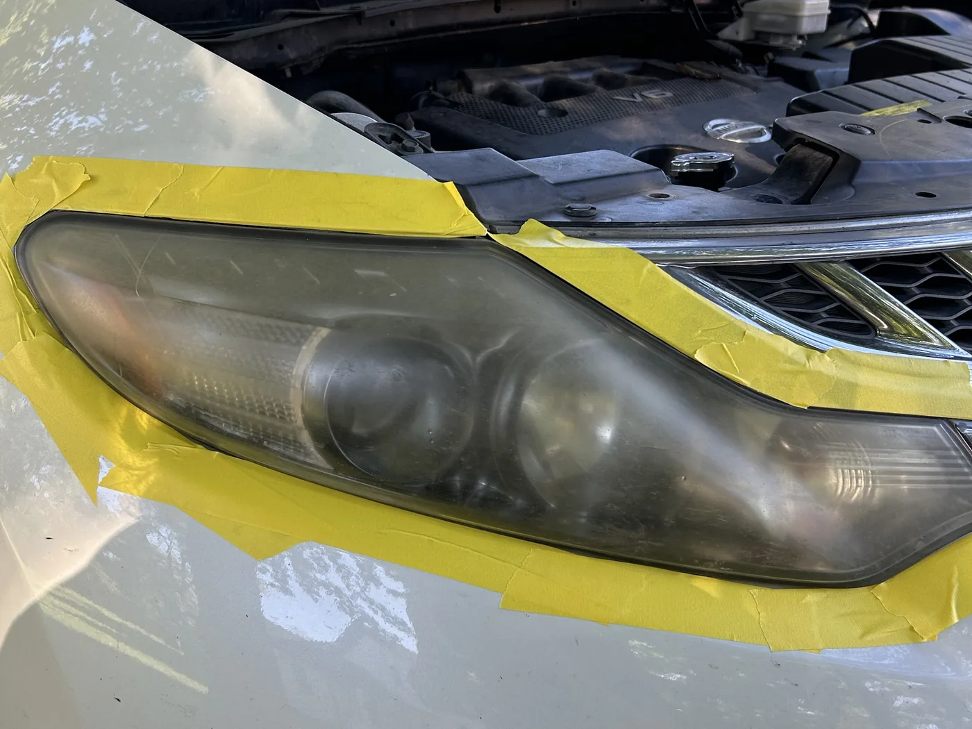 Driver-side headlight on a white Nissan Murano masked off with tape before restoration, the lens fogged over so the projector bowls read as dull grey shapes