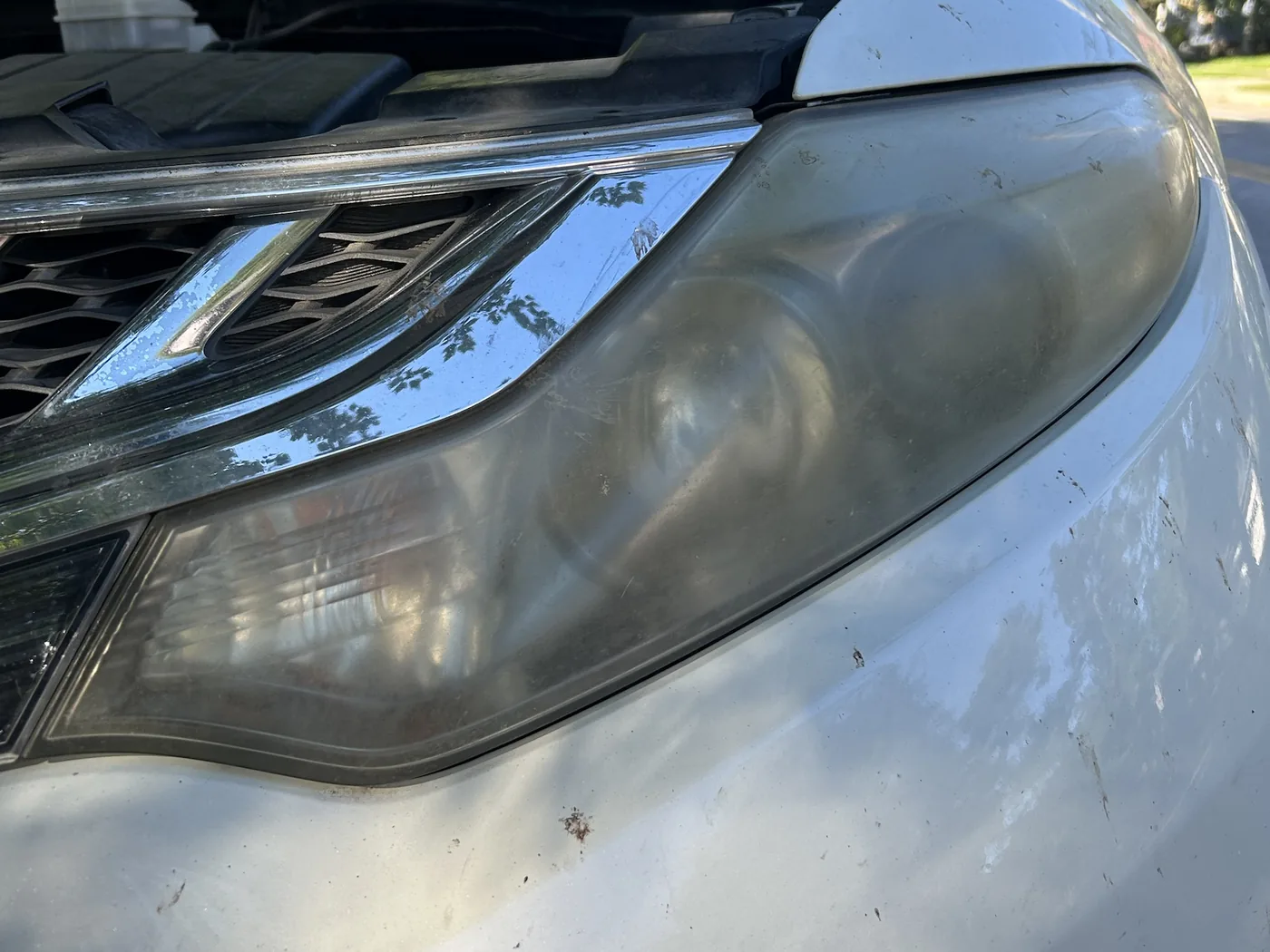 Passenger-side headlight on a white Nissan Murano before restoration, the lens hazed a flat grey so the projector bowls are barely readable