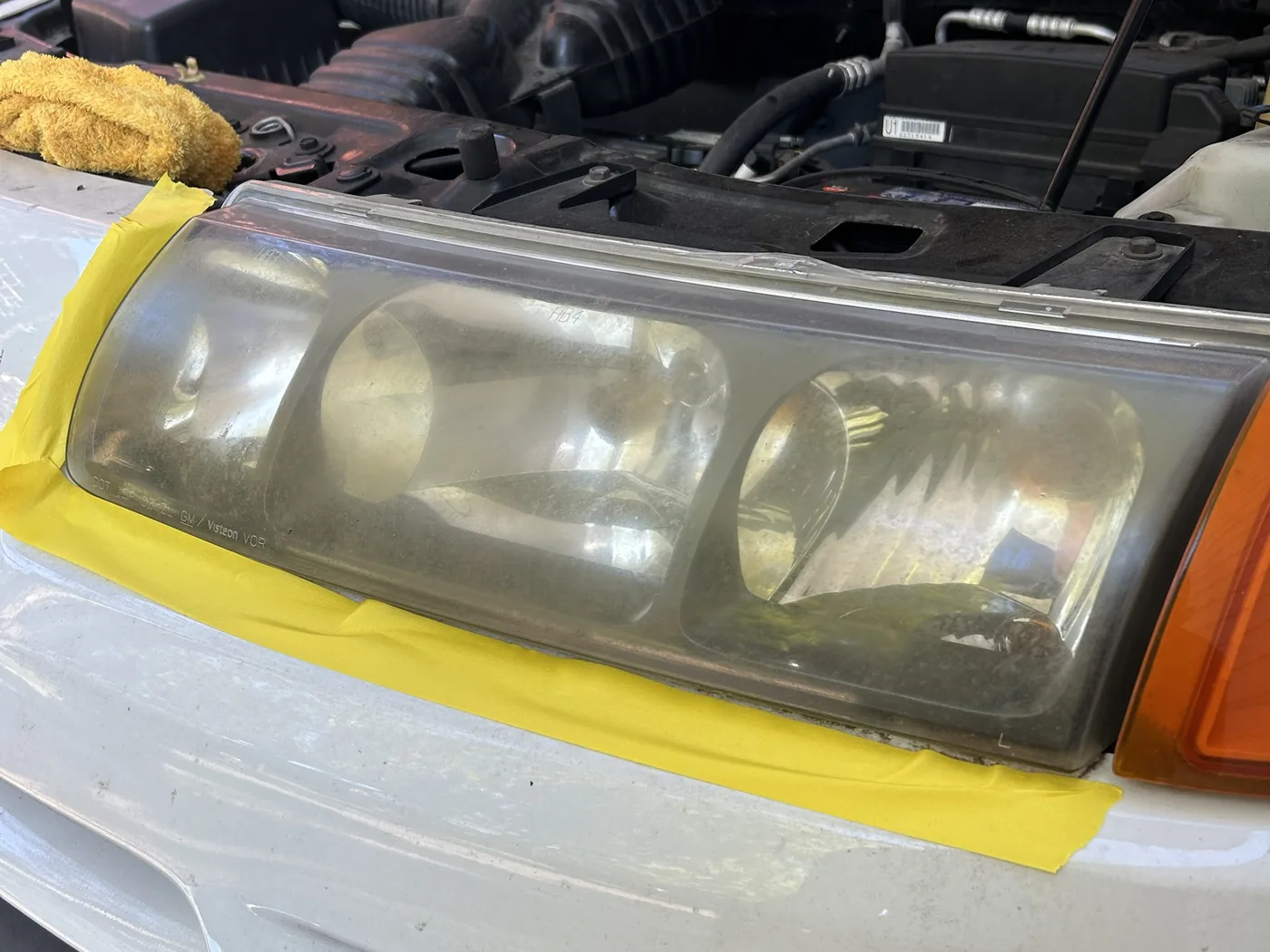 Driver-side headlight on a white Saturn VUE masked off with tape before restoration, the lens deeply yellowed and clouded so the reflectors inside are almost invisible
